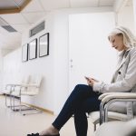 Side view of woman using smart phone while sitting on chair in hospital corridor