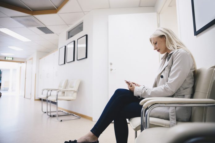 Side view of woman using smart phone while sitting on chair in hospital corridor